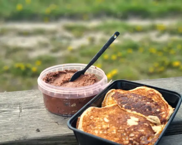 Färdiga proteinpannkakor i en svart behållare bredvid en burk med chokladpålägg, redo att avnjutas utomhus.