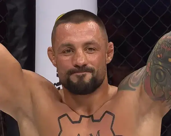 Mateusz Janur, wojownik MMA, z podniesionymi rękami i tatuażami na ramionach i klatce piersiowej, świętuje zwycięstwo.