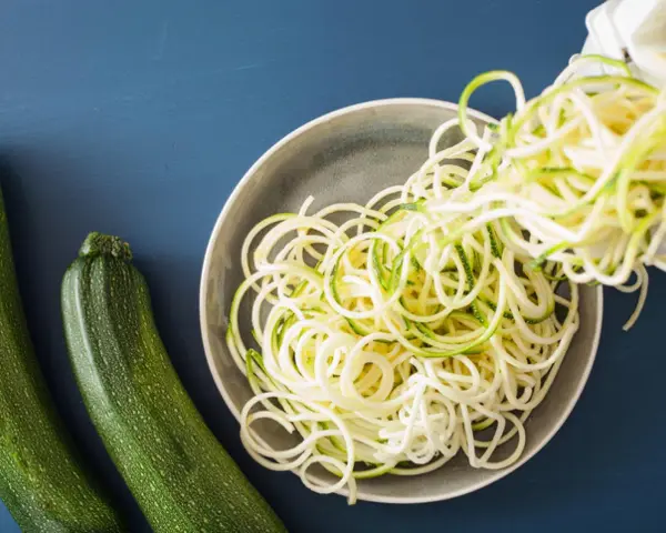 Espaguetis con calabacín recién hechos, listos para servir. Dos calabacines verdes reposan al lado de un plato lleno de fideos de calabacín.