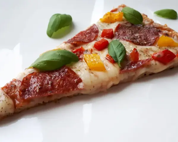 Kawałek pysznej pizzy keto z salami, papryką i bazylią na białym talerzu.