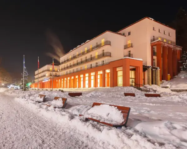 Zimowy wieczór w sanatorium godnym polecenia. Ośnieżone ławki i oświetlony budynek zapraszają do odpoczynku.