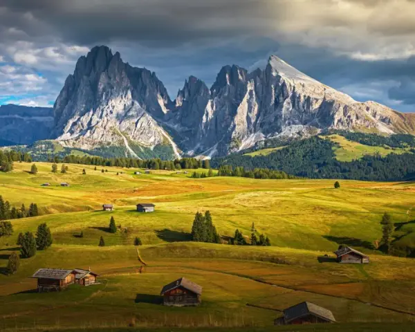 Un paesaggio alpino con un sasso lungo e imponente che domina un sasso piatto e verdeggiante, punteggiato da casette di legno.