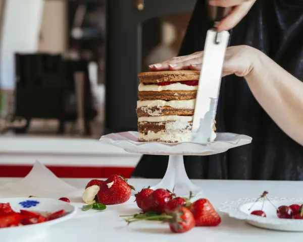 Kobieta wygładza krem na piętrowym torcie, który jest przygotowywany z użyciem pysznego tynku do tortu. Wokół leżą świeże truskawki i wiśnie.