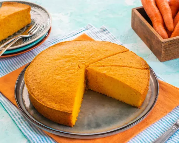 Una fetta di torta di carote sofficissima, appena tagliata da una torta intera su un piatto, con carote fresche in un cesto sullo sfondo.