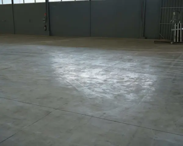 Vaste entrepôt avec une dalle béton qui part en poussière, marquée par des traces de passage.