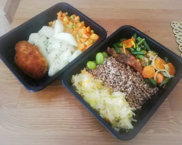 Dwa czarne pojemniki z jedzeniem: kotlet, ziemniaki, warzywa, kasza gryczana, surówka i winogrona.