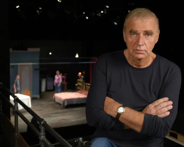 Dawid Głowacki, reżyser teatralny, z rękami skrzyżowanymi na piersi, na tle sceny z łóżkiem i ludźmi.