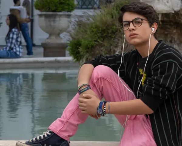 Il ragazzo dai pantaloni rosa, con occhiali e auricolari, ascolta musica seduto vicino a una fontana, mentre altre persone chiacchierano sullo sfondo.