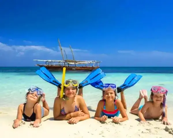 Cztery uśmiechnięte dzieci w maskach do snorkelingu i płetwach leżą na plaży. W tle turkusowa woda i łódź. Idealny dzień, kiedy na Zanzibar!