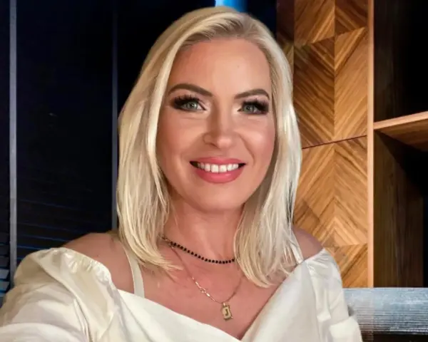 Uśmiechnięta Joanna Janikowska-Samojłowicz w białej bluzce z odkrytymi ramionami, z naszyjnikami na szyi.