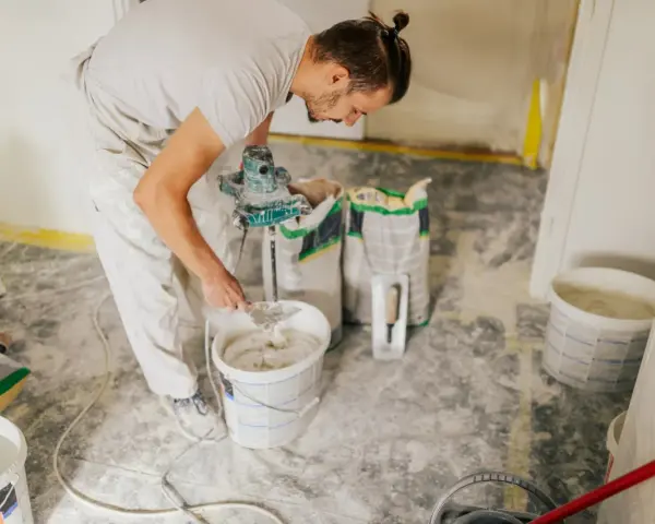 Un ouvrier prépare de l'enduit béton cellulaire dans un seau avec un mélangeur. Des sacs de poudre sont à proximité.