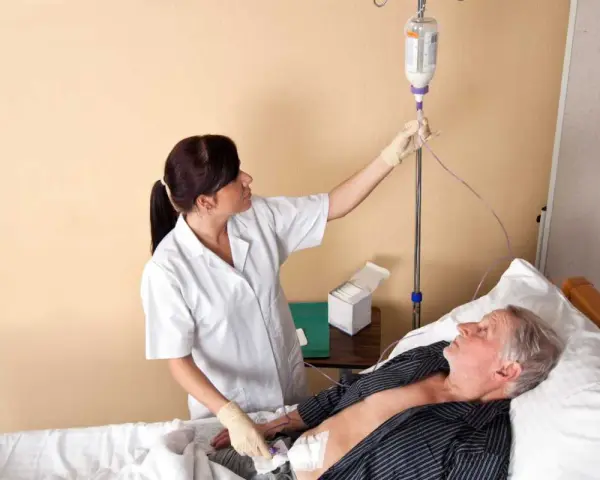 Pielęgniarka podłącza kroplówkę do pacjenta leżącego w łóżku. Widać rurkę od PEG.