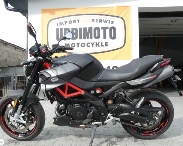 Czarny motocykl Aprilia Shiver 900 z czerwonym stelażem, zaparkowany przed salonem motocyklowym.