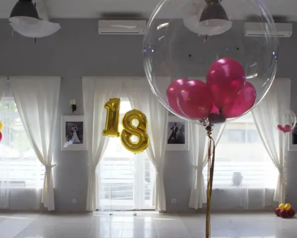 Złote balony z cyfrą 18 i przezroczyste balony z różowymi w środku ozdabiają salę. Balony na stołach tworzą radosną atmosferę.