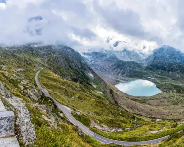 Strada tortuosa sul Passo San Lugano, con lago alpino azzurro e montagne nebbiose sullo sfondo.