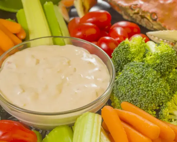 Un bol de sauce crudités crème fraîche entouré de carottes, céleri, tomates cerises et brocolis. Un crabe cuit est visible en arrière-plan.