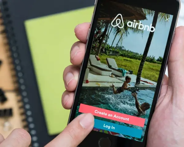 Dłoń trzyma smartfon z aplikacją Airbnb. Czy Airbnb jest bezpieczne? Na ekranie widać basen i dzieci, przyciski "Create an Account" i "Log In".