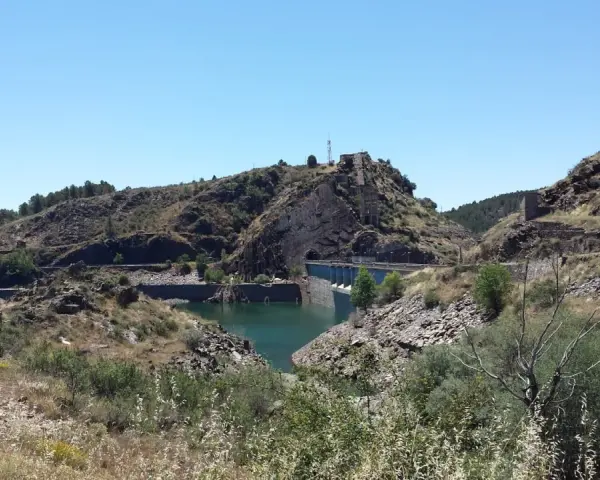 El embalse del Vado, con su presa de hormigón y aguas turquesas, se abre paso entre colinas rocosas y vegetación seca bajo un cielo azul.