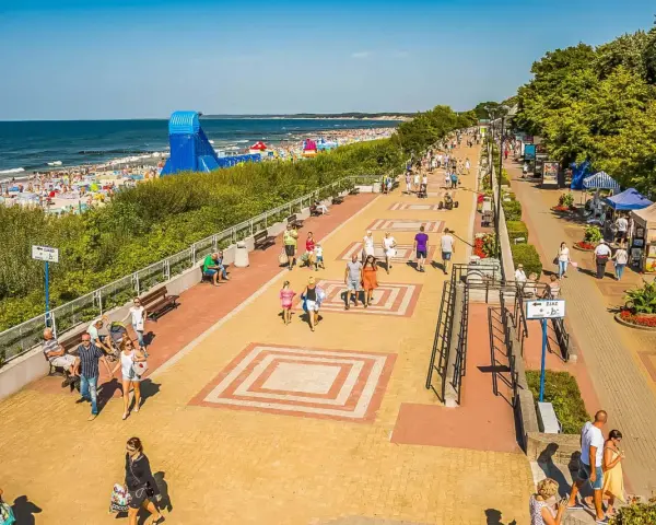 Tętniąca życiem ustka promenada w słoneczny dzień. Ludzie spacerują, odpoczywają na ławkach, a w tle widać plażę pełną turystów i morze.