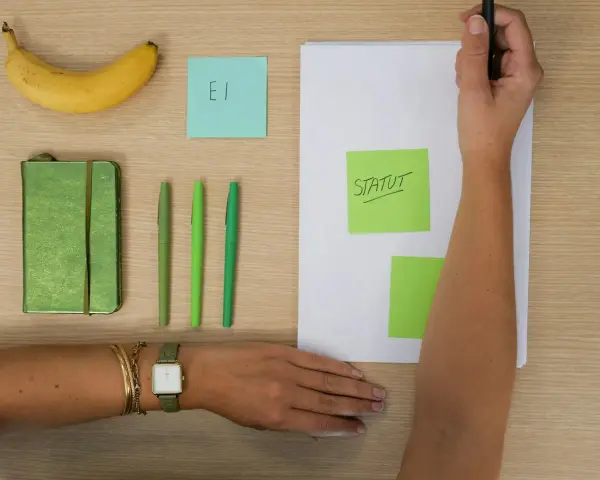 Une main écrit "STATUT" sur un post-it vert, à côté d'un carnet, de stylos et d'une banane. L'acronyme "EI" sur un post-it bleu suggère une **entreprise individuelle**.