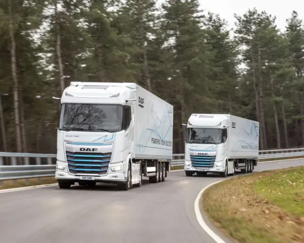 Dwa białe elektryczne DAF XG Plus z naczepami jadące po krętej drodze wśród drzew.