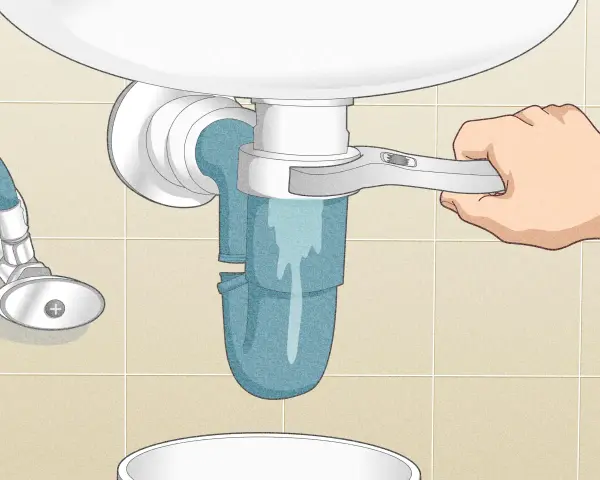 A hand uses a wrench to loosen a sink trap, a step in how to clean a sink drain. Water drains into a bucket below.