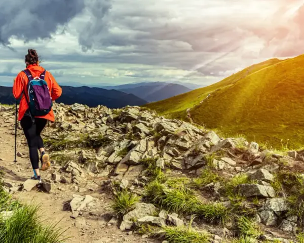 Kobieta w pomarańczowej kurtce wyrusza na szlak. To prawdziwy hiking po polsku, z pięknymi widokami i kamienistą ścieżką.