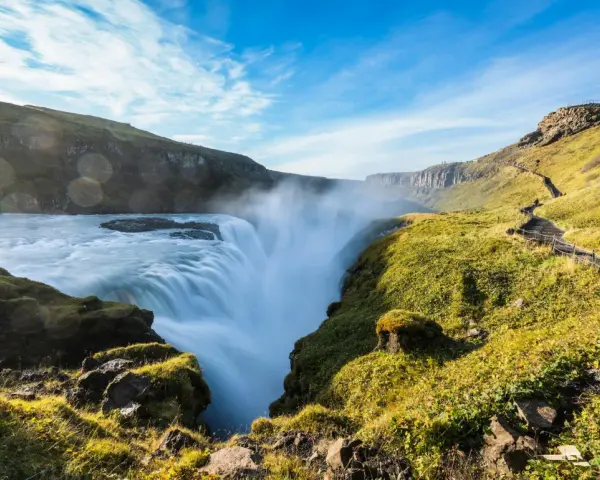 Potężny wodospad Gullfoss w Islandii, z mgiełką unoszącą się nad kaskadami. Ścieżka turystyczna wije się po zielonym zboczu.