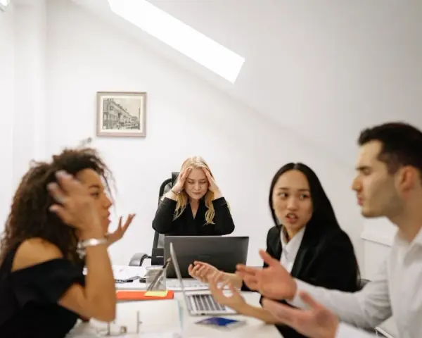 Un gruppo di colleghi di lavoro discute animatamente. Una donna bionda sembra stressata, tenendosi la testa tra le mani.
