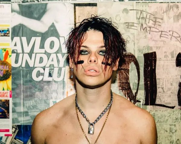 Yungblud, "The Emperor", z charakterystycznym makijażem i łańcuchami, na tle plakatów.