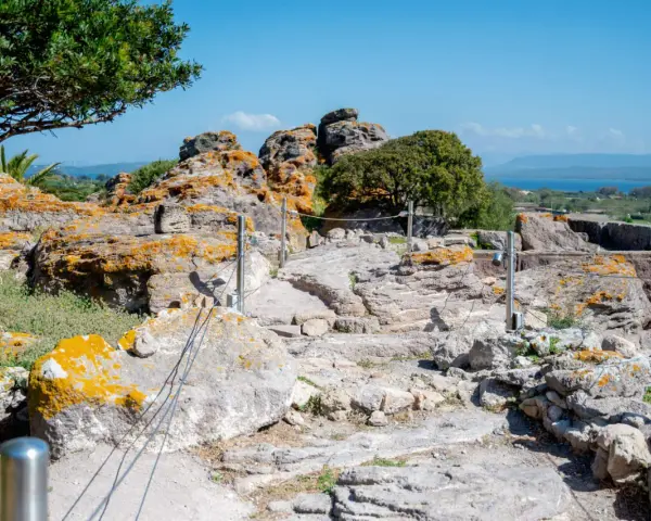 Ancient Italian ruins with weathered stones covered in orange lichen, a pathway winding through the site, and a distant blue sea under a clear sky.