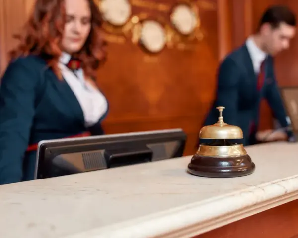 Recepcjonistka i recepcjonista za ladą hotelową. Dzwonek do obsługi gości, który pomoże dowiedzieć się, ile kosztuje doba hotelowa.