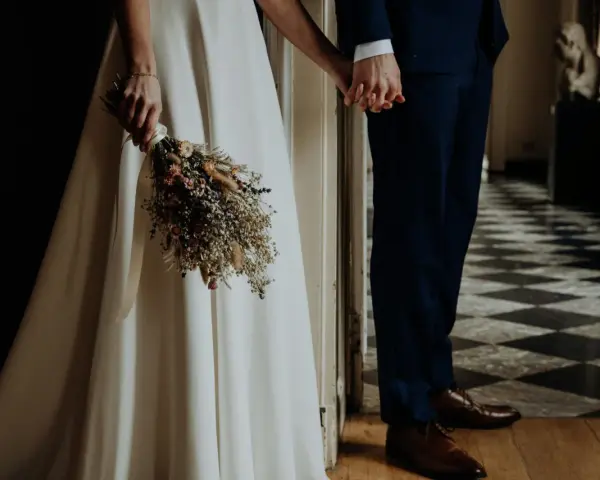 Para młoda trzyma się za ręce, ona z bukietem suszonych kwiatów. Wesele motywy w eleganckim wnętrzu.