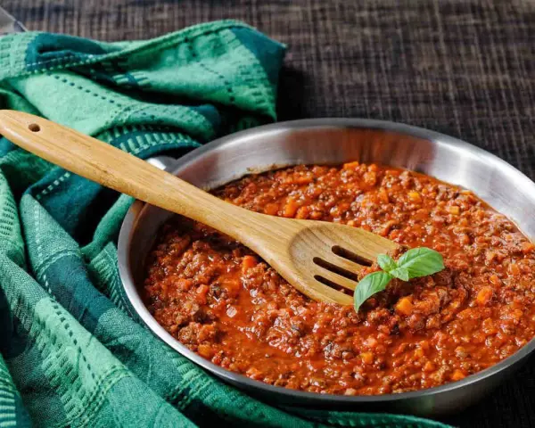 Un saporito ragù di cinghiale in padella, pronto per condire la pasta. Un cucchiaio di legno e una fogliolina di basilico completano la scena.