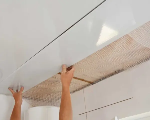 Pose de lambris PVC au plafond avec clips. Les mains ajustent une lame blanche brillante sur une structure, révélant un treillis blanc en dessous.