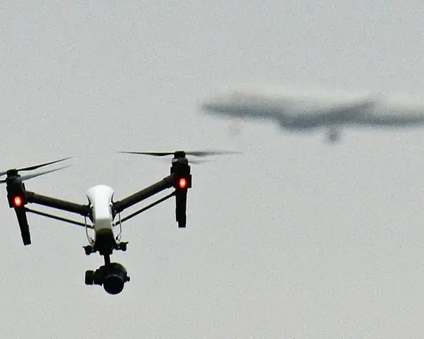 A white drone with red lights hovers in the sky, possibly on a security mission, as a blurred airplane flies in the distance.