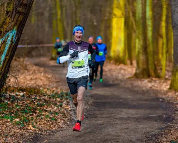 Uśmiechnięty biegacz z numerem 4 na koszulce, w niebieskiej opasce, biegnie leśną ścieżką. To bart przedwojewski, który pokonuje trasę biegu.