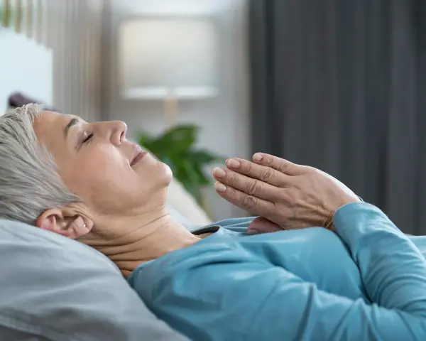 Frau liegt entspannt im Bett, Hände vor der Brust gefaltet. Sie praktiziert eine Technik, die wirkt Meditation im Schlaf.