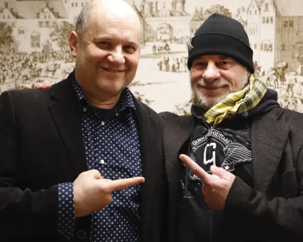 Deux hommes souriants, l'un avec une chemise à pois, l'autre avec un bonnet, se montrent du doigt. Un chanteur de confiance.