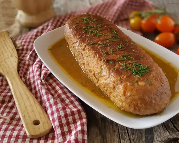 Polpettone in padella dorato, guarnito con erba cipollina, servito su un piatto bianco con salsa. Accanto, una spatola di legno e pomodorini.