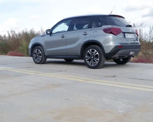 Szary Suzuki Vitara Hybrid z czarnym dachem stoi na betonowej nawierzchni z żółtymi pasami.