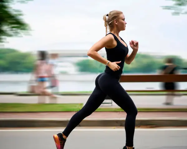 Donna bionda in abbigliamento sportivo nero corre con una postura corsa perfetta lungo un percorso alberato.