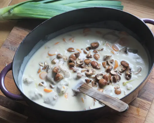 Un plat de blanquette de veau facile mijote dans une cocotte, garni de champignons et de carottes. Un poireau est posé à côté.
