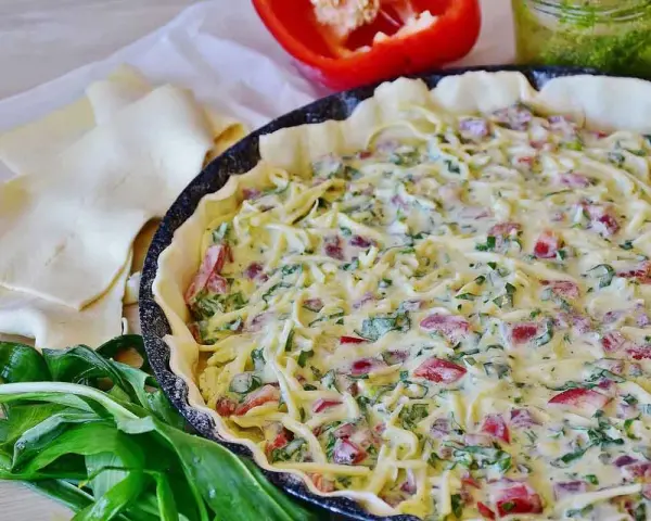 Tarte salée garnie de fromage, poivrons et ail des ours, prête à cuire. Pâte feuilletée et poivron rouge à côté.