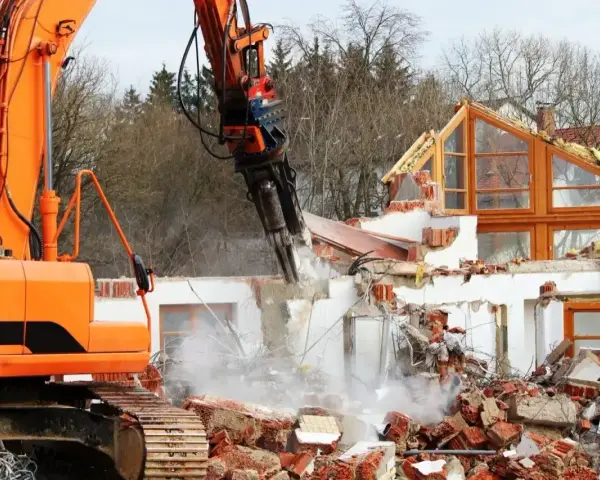 Pomarańczowa koparka niszczy dom, unosząc tumany kurzu i gruzu.