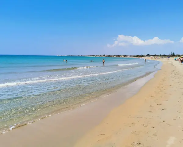 A wide, sandy beach with clear turquoise water and a few people swimming and sunbathing under a bright blue sky.