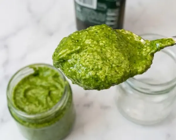 Cucchiaio pieno di pesto senza pinoli, cremoso e verde brillante, accanto a un barattolo.