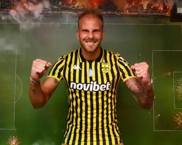 Uros Racic, con su nueva camiseta a rayas amarillas y negras, celebra con los puños en alto. El fondo simula un campo de fútbol con bengalas.