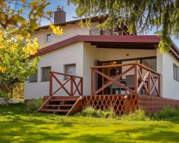 Bungalow z drewnianym tarasem i schodkami, otoczony zielonym ogrodem i kwitnącym drzewem.