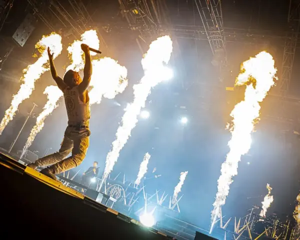 Un chanteur de groupe hard rock des années 2000, micro en main, saute sur scène entouré de jets de flammes spectaculaires.